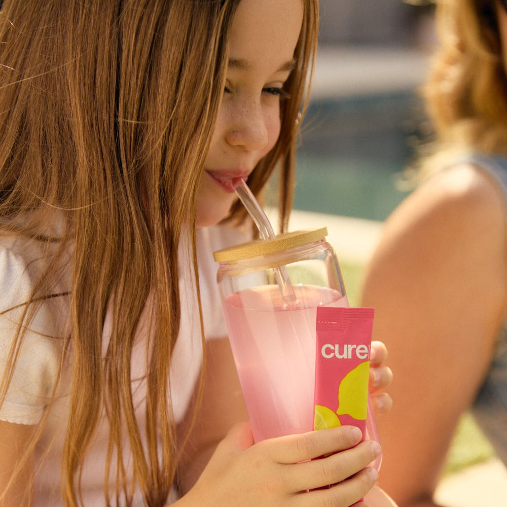 Young girl sipping a pink sports drink from a glass jar with a straw while holding a pink packet labeled cure outdoors