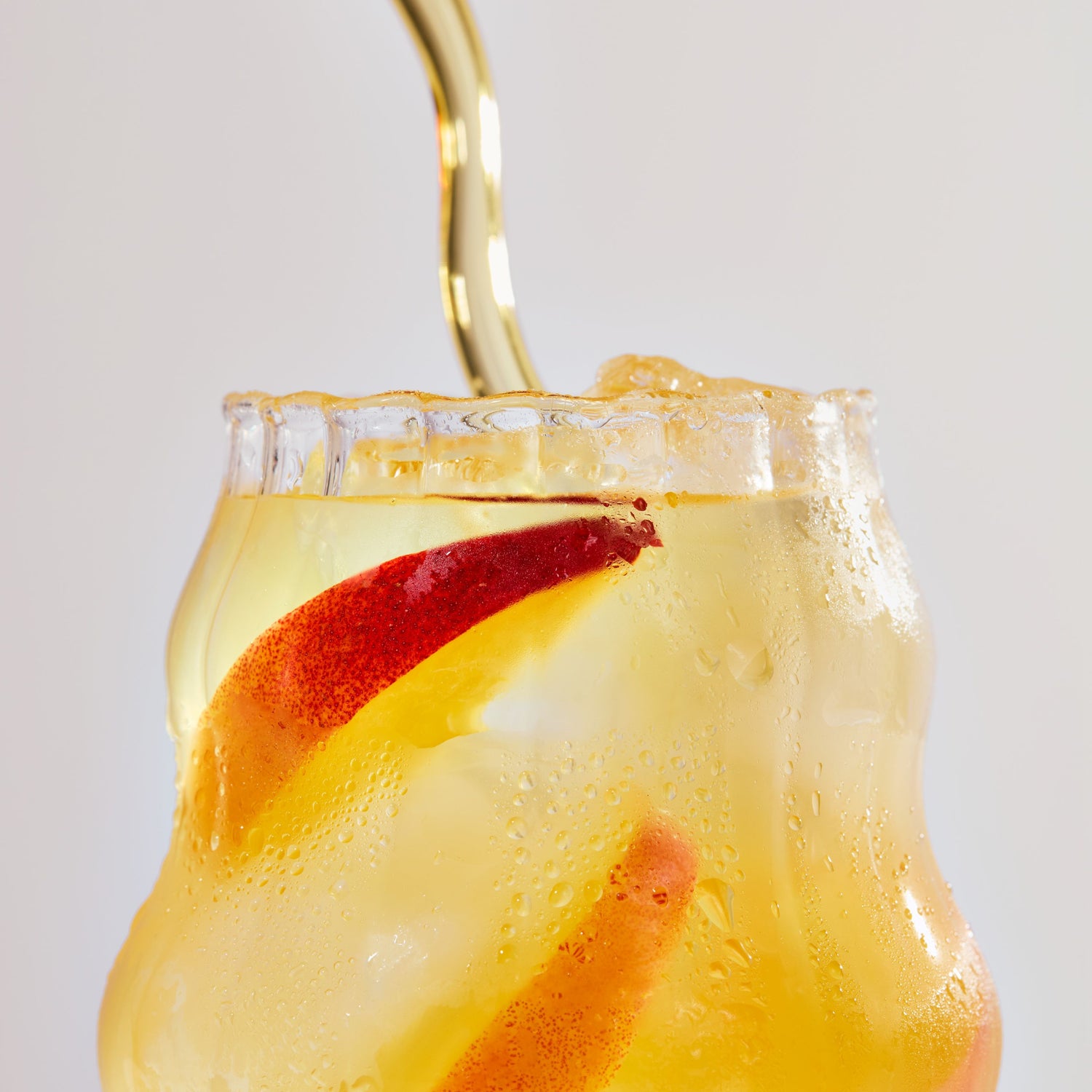 Peach and mango iced cocktail with peach slices, ice, and condensation in a curvy glass