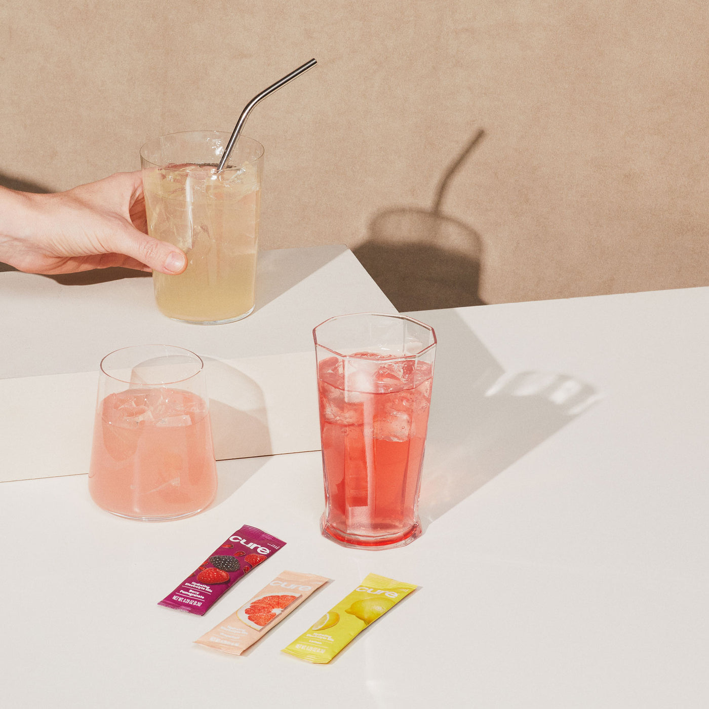 Hand holding yellow beverage, two more glasses (pink, red) with ice; 'Cure' packets in berry, grapefruit, lemonade, on a warm brown background.