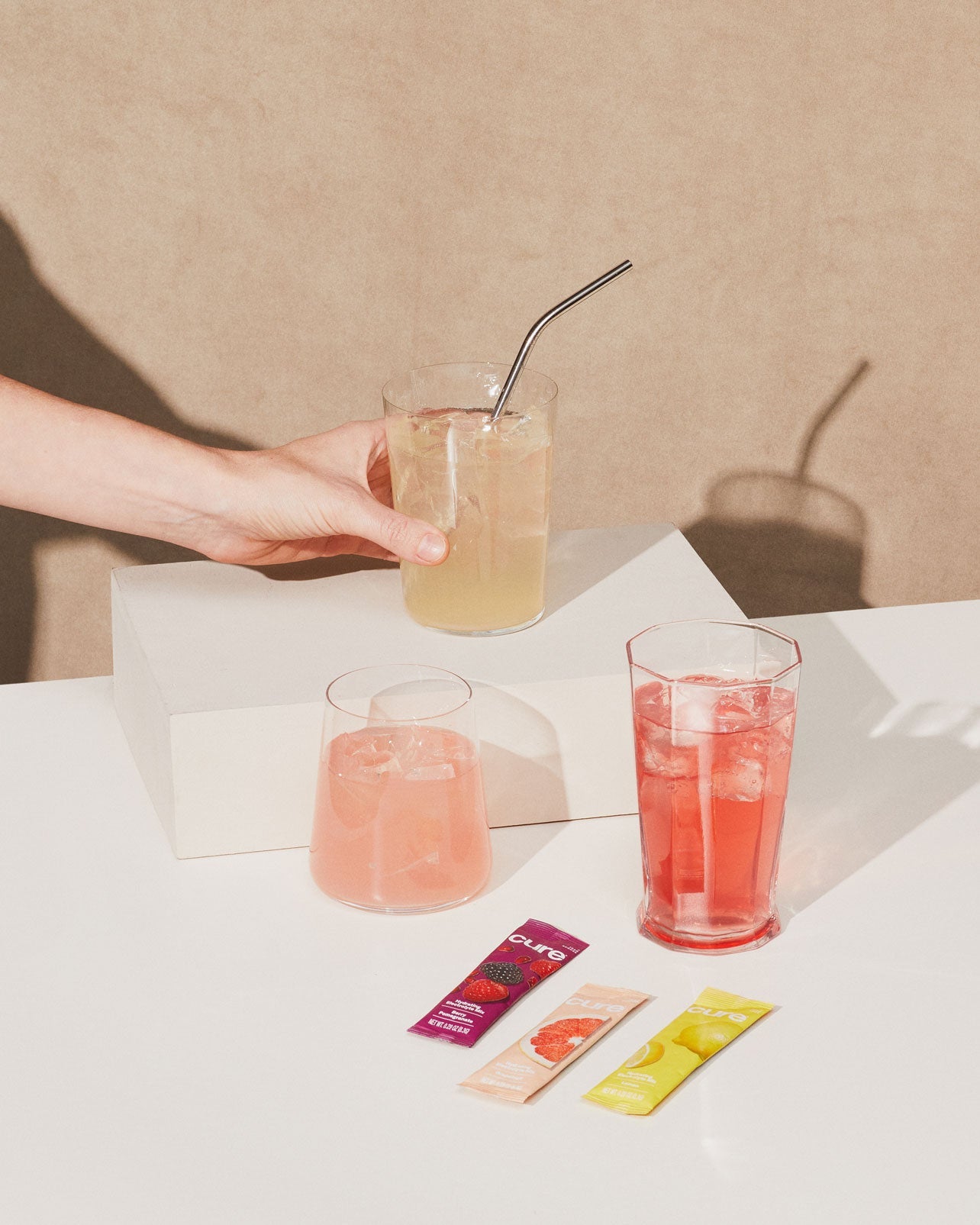 Hand holding yellow beverage, two more glasses (pink, red) with ice; 'Cure' packets in berry, grapefruit, lemonade, on a warm brown background.