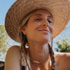 Woman with braided hair in a large straw hat, smiling in a sunny outdoor setting.