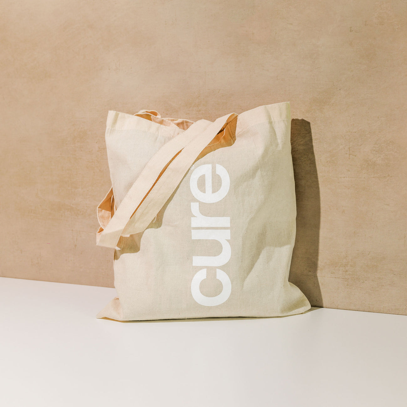 Front view of Tote Bag showing vertical white cure logo, natural cotton, 24in handles, 15.5 x 15 in on beige background