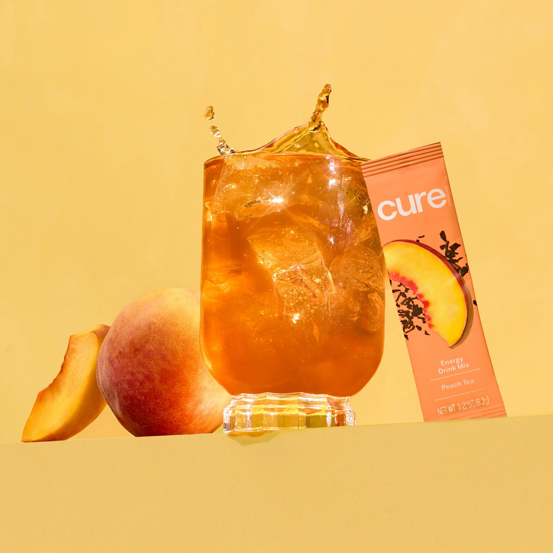 Iced peach tea in a glass with a peach and a tilted packet labeled cure; text: 'cure', 'Energy Drink Mix', 'Peach Tea', 'NET WT 0.22 OZ (6.2g)'