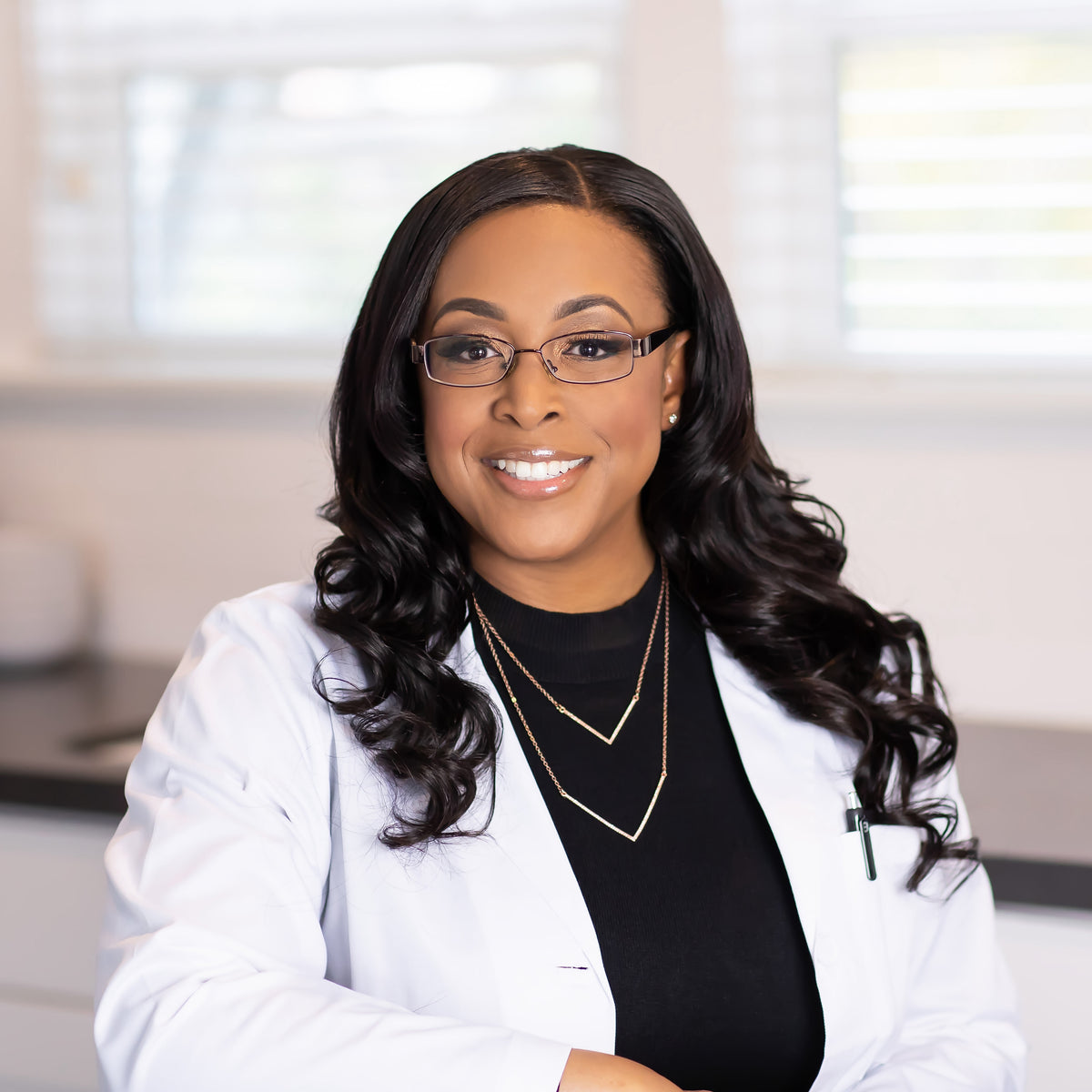 Portrait of a smiling woman with black hair, glasses, white lab coat, in a bright interior.