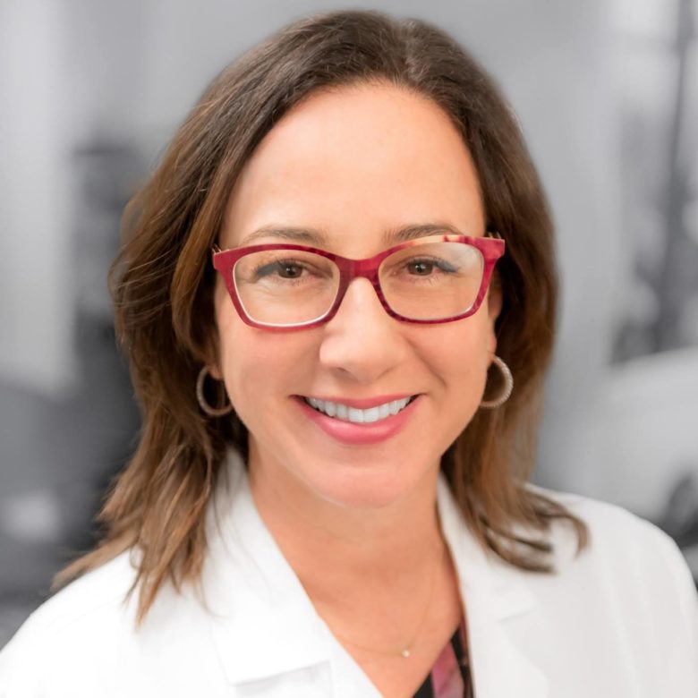 Smiling healthcare worker with dark hair, red glasses, in white lab coat, exuding confidence.