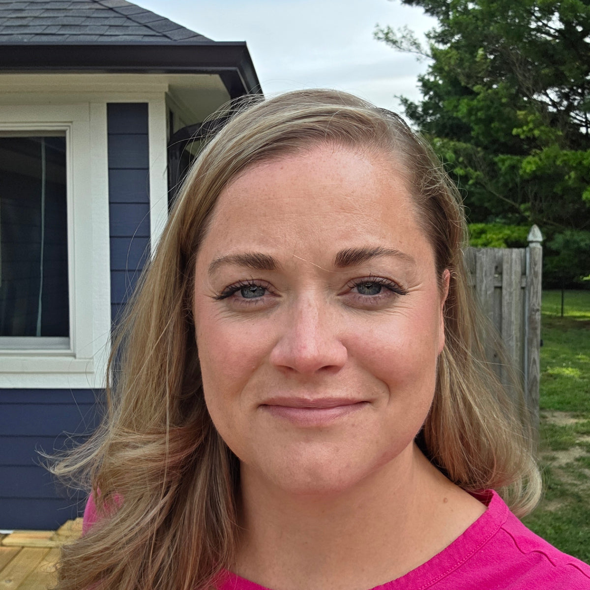 Close-up of a smiling woman with blonde hair standing outside near a blue house and wooden fence.