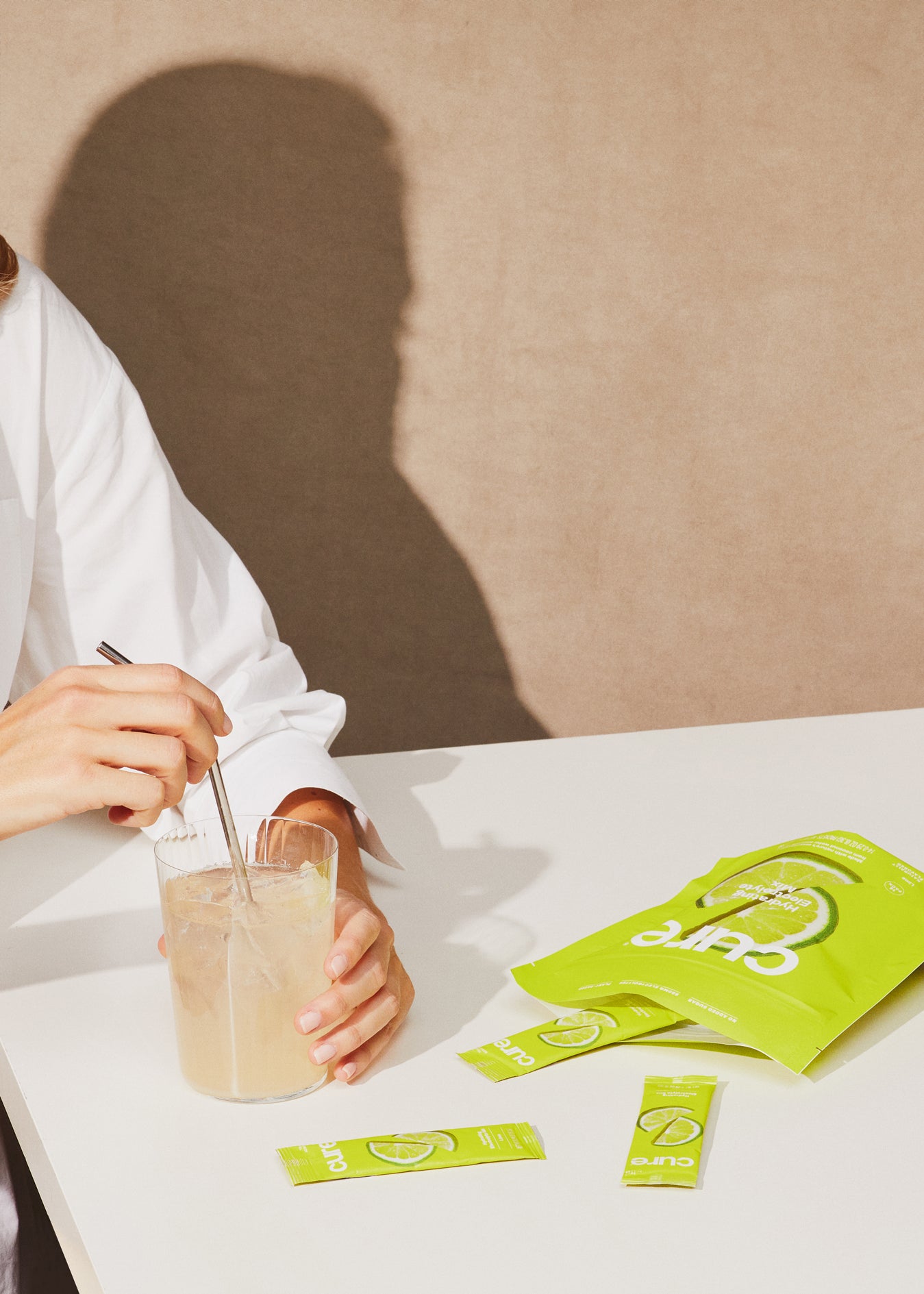 Person stirs light drink with 'Cure' packets on white table, beige background.