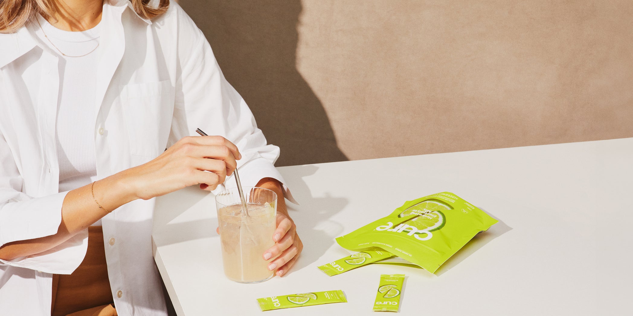 Person stirs light drink with 'Cure' packets on white table, beige background.