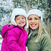 Smiling adult and child in white knit hats and winter clothes with snowy trees in the background.