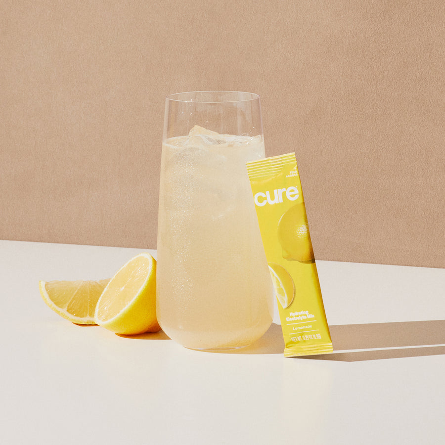 Glass of lemonade with 'cure' mix packet and lemon slices on white surface.