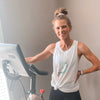 Fit woman with hair in a bun, in a white tank top by an exercise bike, suggesting a workout.