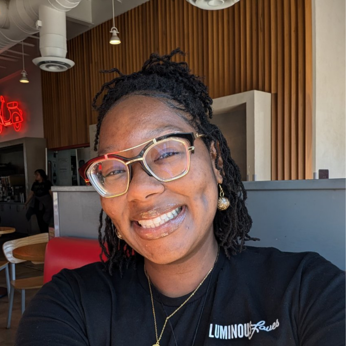 Smiling person with glasses and earrings in a cafe with wooden paneling and red seating.