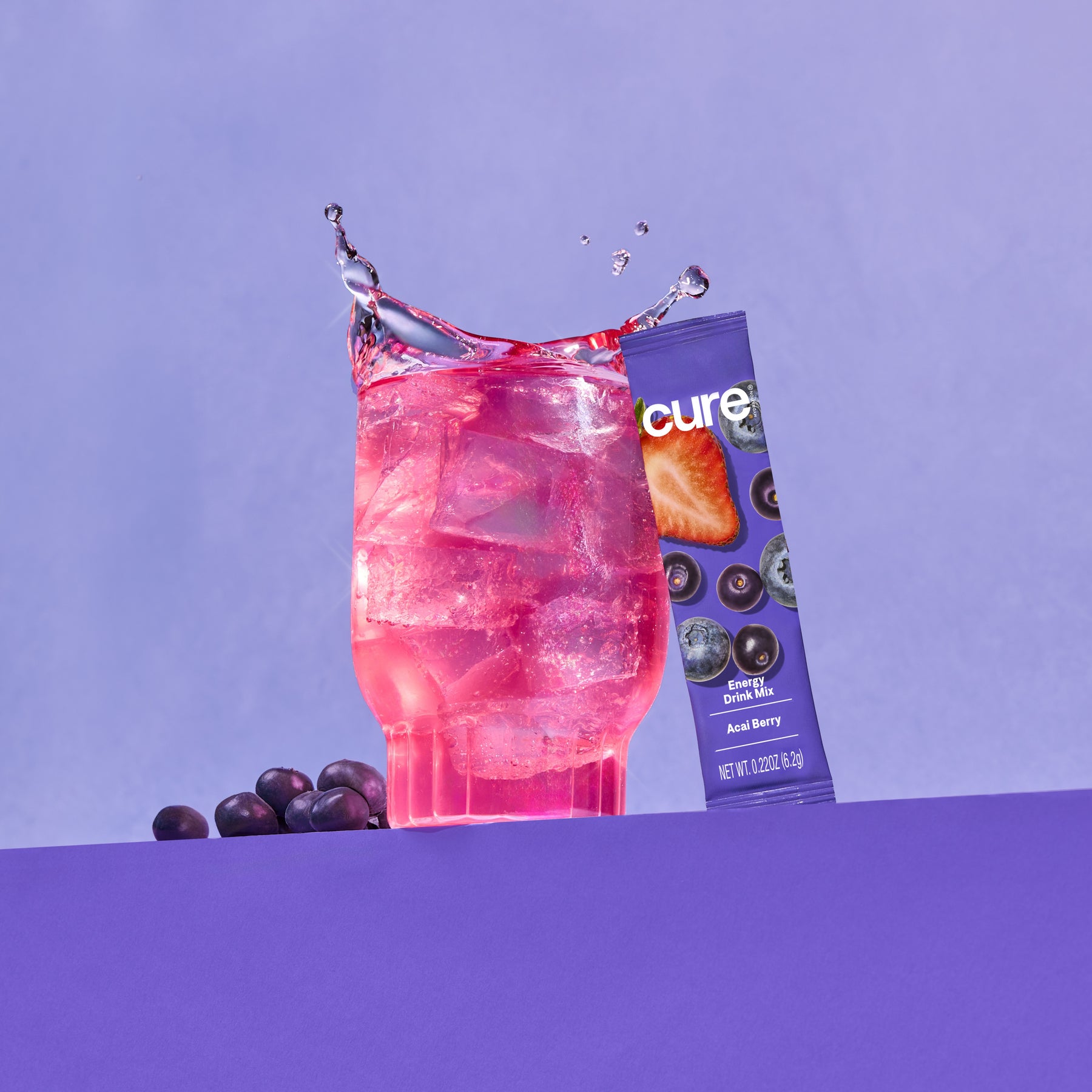 Pink iced drink splashing in glass beside Acai Berry Energy Drink Mix packet labeled 'cure' — NET WT. 0.22OZ (6.2g)