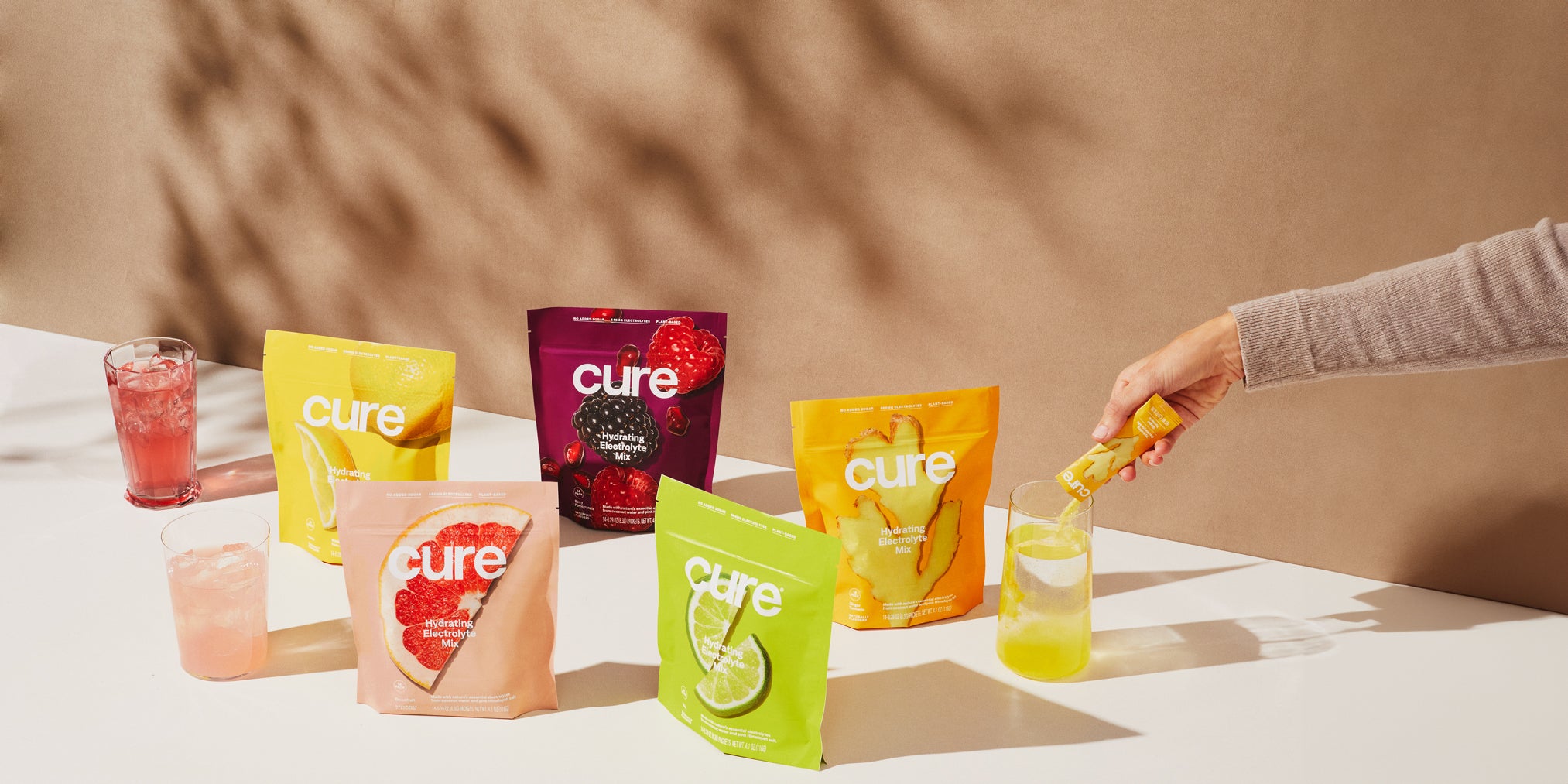 'Cure' electrolyte packets and drinks with a hand pouring a yellow mix, neutral backdrop.