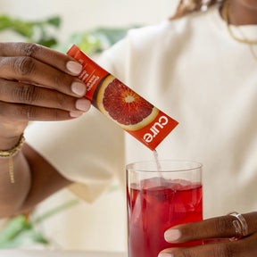 Person pouring Cure Blood Orange electrolyte mix packet into a glass of iced red drink