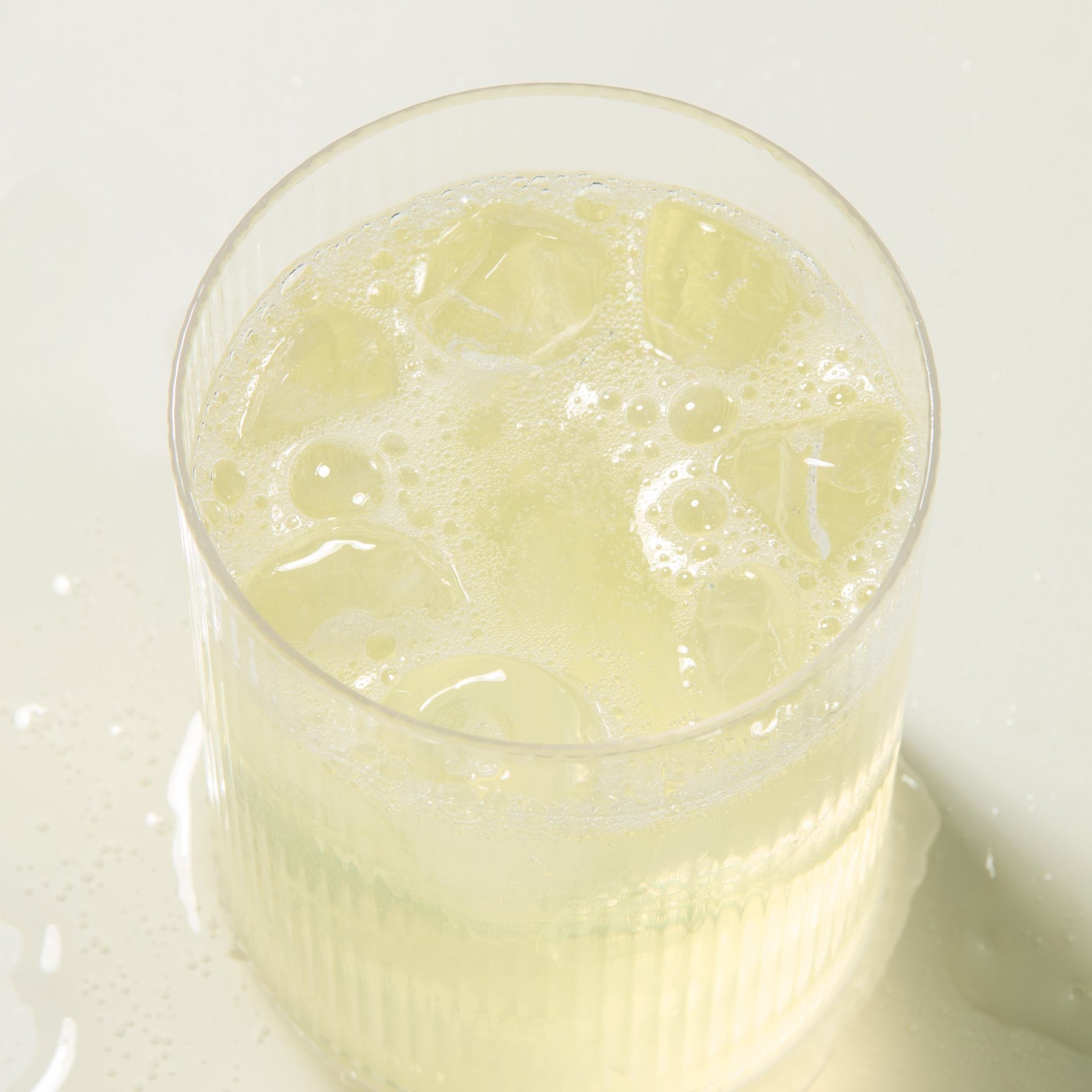 Top-down view of Lemonade in a ribbed glass showing ice cubes and light foam on a pale tabletop