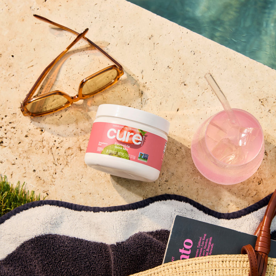 Angled top view of Strawberry Kiwi electrolyte mix jar showing pink Cure label, poolside with sunglasses and pink iced drink