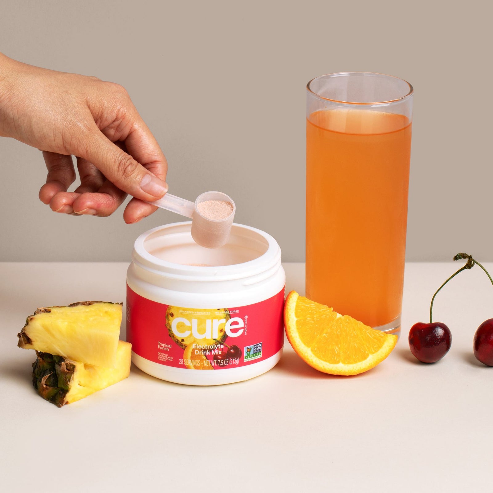 Close-up front view of Tropical Punch bulk jar showing a scoop of powder and a glass of orange drink with pineapple and orange slices on a beige background
